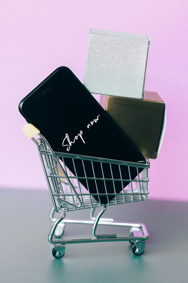 A close-up of a miniature shopping cart containing a black phone that reads “Shop now.”