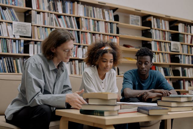 Three students sitting in a university library looking at a stack of books. 
