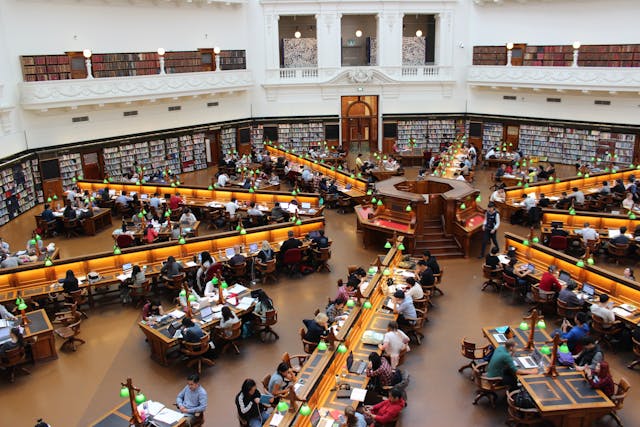 A large group of college students studying inside a huge, well-lit university library. 
